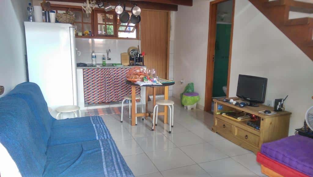 Sala de estar do airbnb Mi Casa Su Casa Lofts. No canto esquerdo está o sofá da sala, em sua frente há um pequeno hack com uma TV em cima, no centro do cômodo há uma mesa redonda com dois banquinhos, no canto esquerdo há uma geladeira e no fundo está a pia da cozinha.