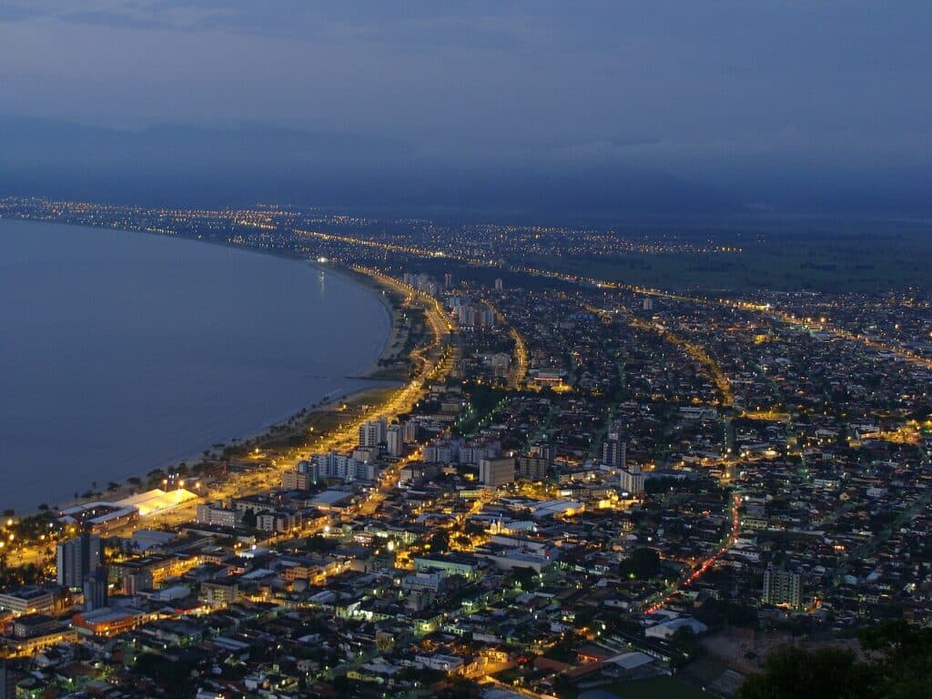 Foto aérea da cidade de Caraguatatuba ao anoitecer. Várias casas e prédios rodeiam a costa que fica na lateral esquerda da imagem. - Foto: Silvio Emanuel Fernandes via Wikimedia Commons