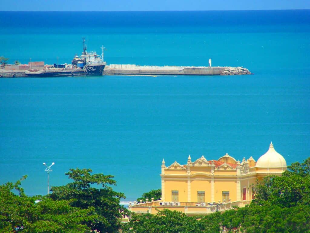 Prédio histórico amarelo, rodeado de árvores verdes, com o mar de fundo e o porto de Maceió. Representa um dos pontos turísticos de Maceió