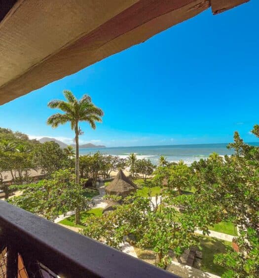 Vista de uma varanda com vista para uma praia tropical, palmeiras, vegetação e um céu azul claro sobre o oceano. O local é o hotel Vista do mar da sacada do Nacional Inn Ubatuba, um dos hotéis em Ubatuba