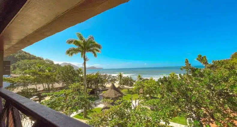 Vista de uma varanda com vista para uma praia tropical, palmeiras, vegetação e um céu azul claro sobre o oceano. O local é o hotel Vista do mar da sacada do Nacional Inn Ubatuba, um dos hotéis em Ubatuba