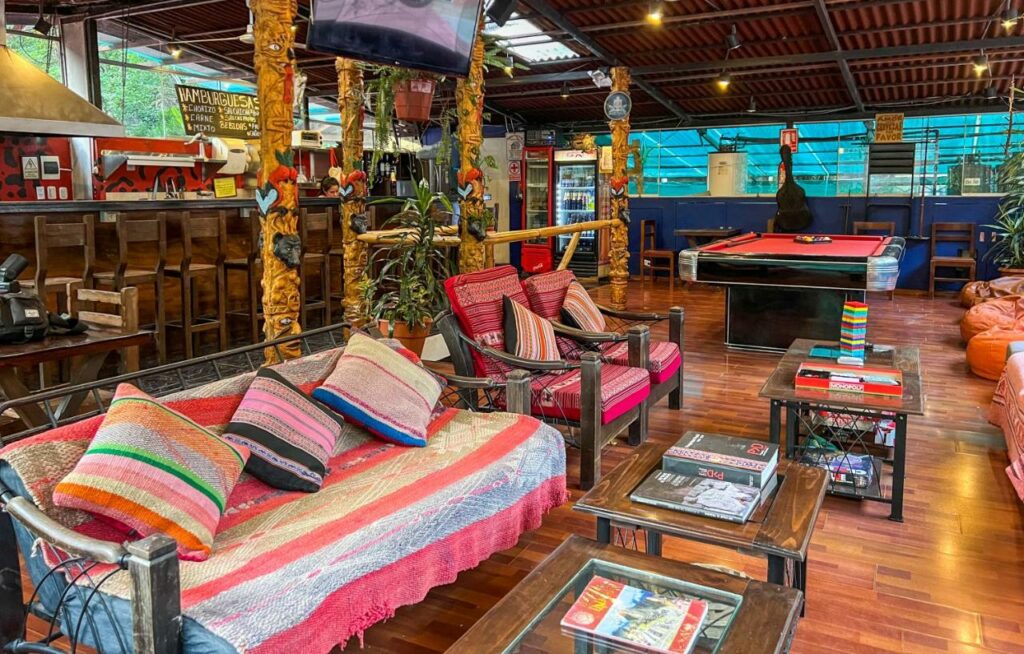 lounge compartilhado do Nativus Hostel Machu Picchu com vários sofás, pufes e cadeiras dispostos de frente uns aos outros, com mesas de centro equipadas com livros e revistas, além de uma mesa de bilhar ao fundo e uma área de bar à esquerda da imagem.