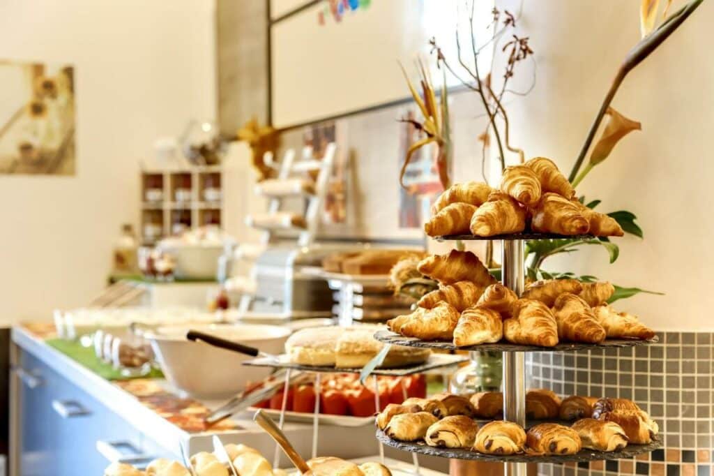 Itens de café da manhã do Novotel Château de Versailles. A foto está focada nas bandejas de croissants e pain au chocolat, mas ao fundo há outros pães, cereais, bolos e diversos alimentos.