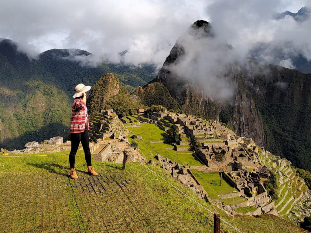 mulher em pé e de costas para a câmera, no lado esquerdo da imagem, usando um chapéu e olhando para a montanha de machupicchu que está parcialmente escondida por algumas nuvens no lado direito da imagem