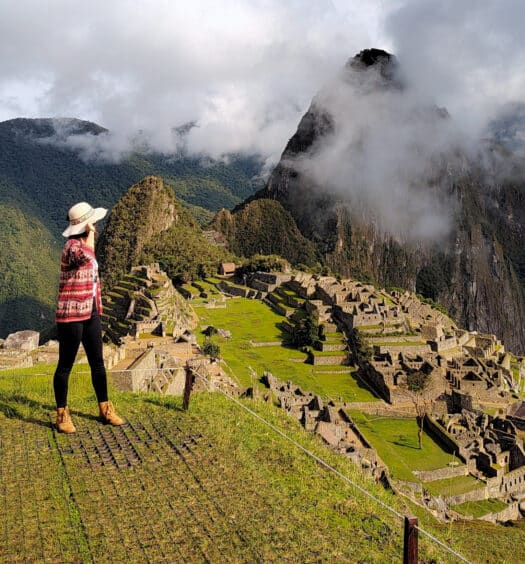 mulher em pé e de costas para a câmera, no lado esquerdo da imagem, usando um chapéu e olhando para a montanha de machupicchu que está parcialmente escondida por algumas nuvens no lado direito da imagem