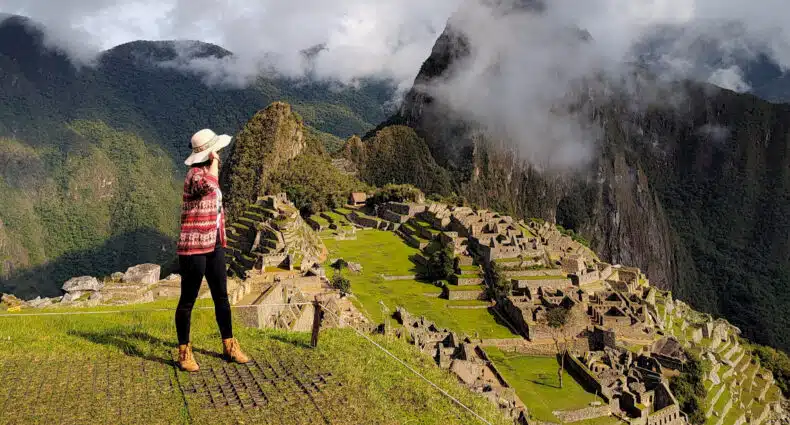 mulher em pé e de costas para a câmera, no lado esquerdo da imagem, usando um chapéu e olhando para a montanha de machupicchu que está parcialmente escondida por algumas nuvens no lado direito da imagem