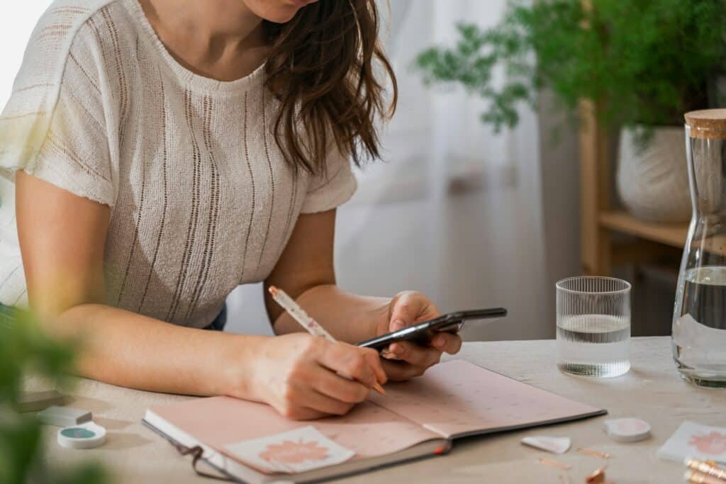 uma mulher escrevendo em um caderno enquanto olha no celular, há água também em cima da mesa, para ilustrar o post de seguro viagem cobre cancelamento de viagem