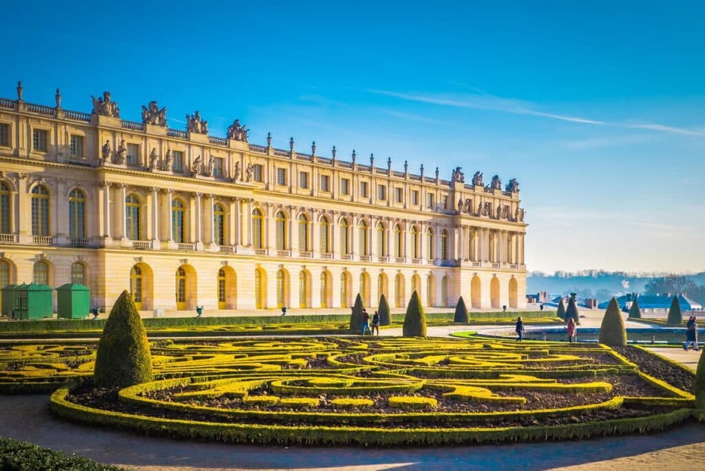 Fachada do Palácio de Versalhes, que é extensa e repleta de janelas. Em frente há um jardim com arbustos e árvores onde algumas pessoas caminham. - Foto: Civitatis