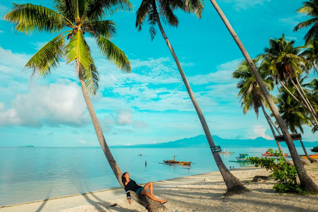 Uma mulher relaxa em uma palmeira inclinada na praia de Paliton, com barcos na água e um céu azul com nuvens dispersas. Ilustra o post de melhores praias nas Filipinas.
