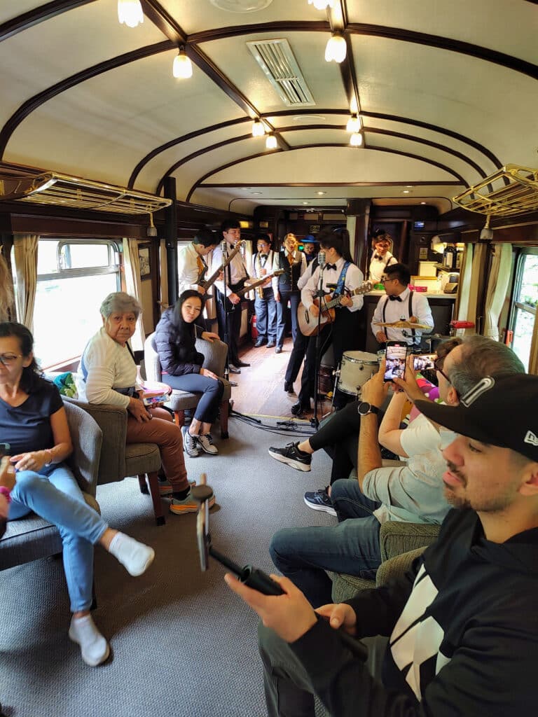 várias pessoas sentadas, mexendo no celular, enquanto há uma banda vestida de roupa social ao fundo, tocando música ao vivo. Há uma guitarra, um baixo e um saxofone.