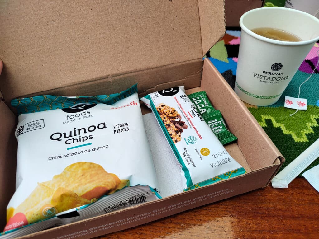 caixa de papelão aberta com um pacote pequeno de aperitivo de quinoa, semelhante aos salgadinho Ruffles, uma barrinha de chocolate e quinoa e duas balinhas de coca. Ao lado da caixa, há um copo de papel com chá. Esse é o lanche fornecido pela perurail