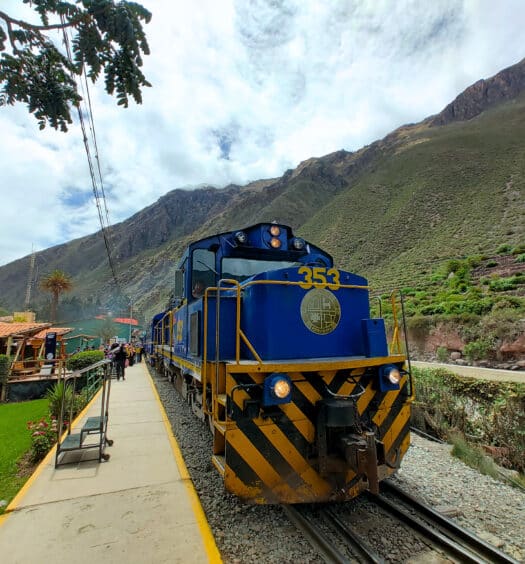 visão panorâmica do trem da Perurail vista de frente para o vagão condutor, parado na estação. Ao redor é possível enxergar as montanhas peruanas e a plataforma de acesso.