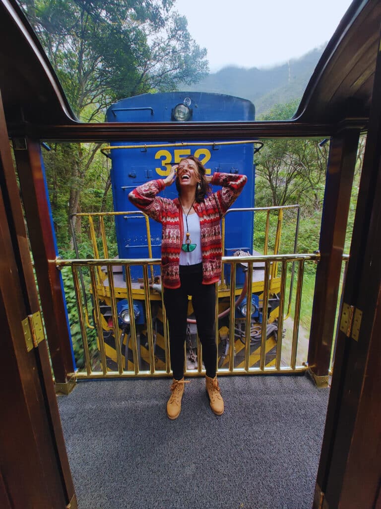 mulher em pé, na parte aberta do vagão vistadome, sorrindo com as mãos segurando os cabelos que estão voando ao ar livre e em seu rosto. Atrás dela é possível ver o vagão condutor na cor azul