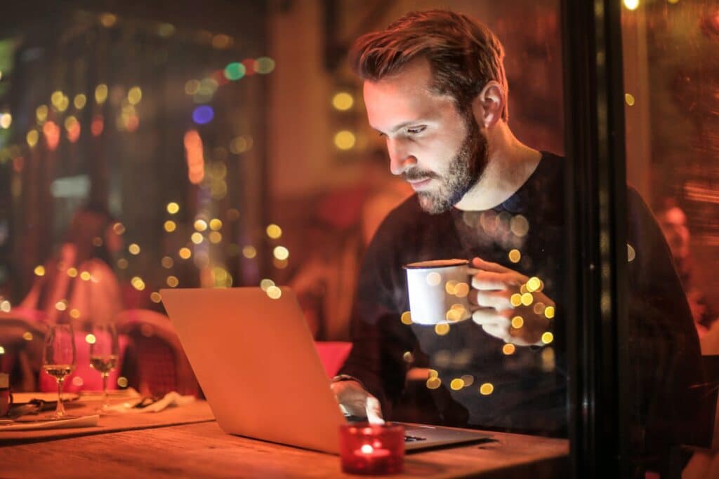 Um homem sentado em uma cafeteria com uma notebook aberto na sua frente, com uma mão ele está digitando, com a outra está segurando uma xícara de café