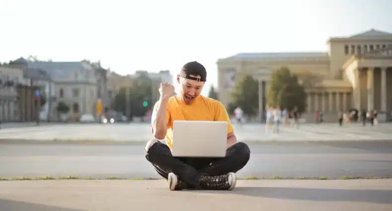 Um homem sentado em um espaço público, com um notebook aberto em seu colo, ele está comemorando e vibrando em frente à tela, para representar cotação de seguro viagem