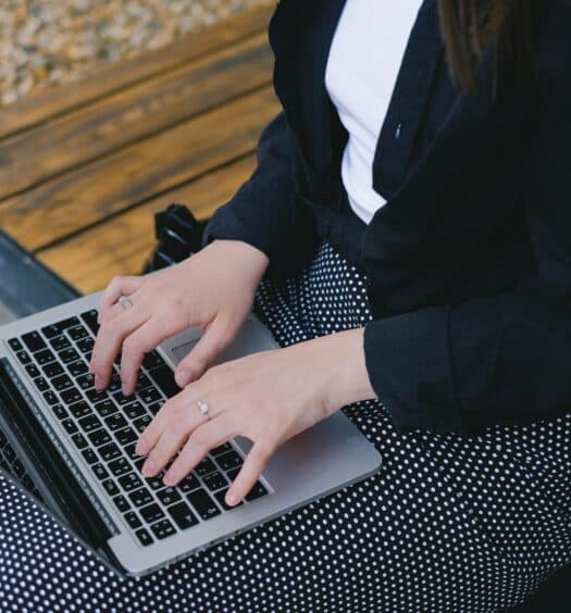 Uma mulher sentada em um banco de madeira com um notebook aberto sob suas pernas enquanto ela digita, para representar contratar seguro viagem é obrigatório