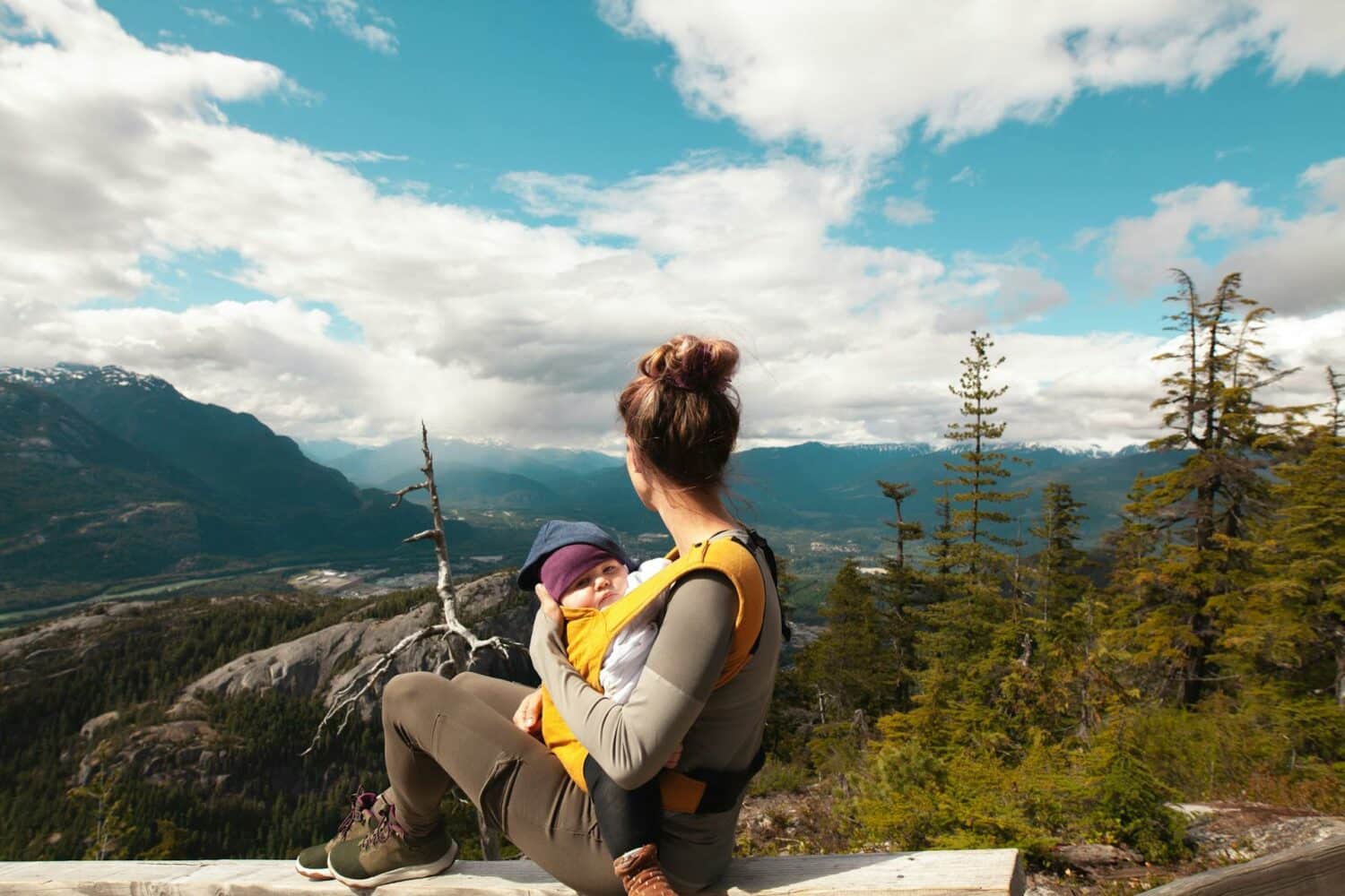 Uma mulher sentada observando montanhas, com muitas árvores ao seu redor, em seu colo está um bebê sendo segurado pelos dois braços da mulher, para representar seguro viagem bebê