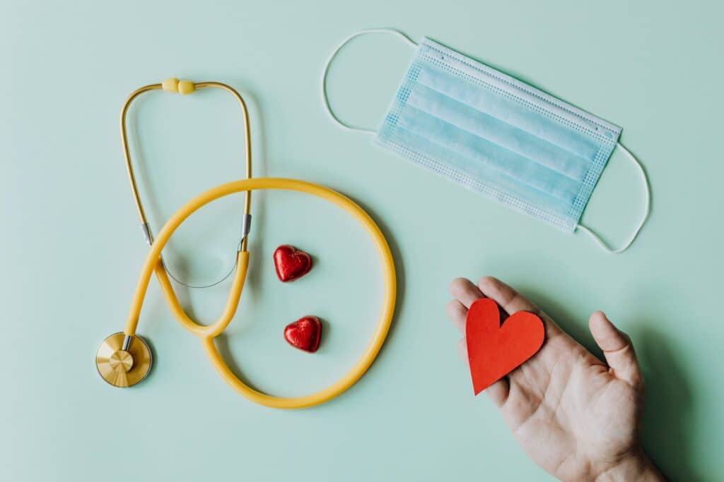 Um estetoscópio, uma máscara descartável e uma mão segurando um coração cortado em papel vermelho, para representar quanto custa atendimento médico no exterior