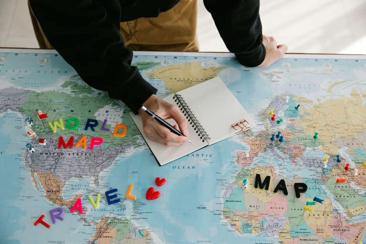 Um mapa mundi aberto com algumas peças que imitam letras formando da palavra Travel em cima dele, há também um caderno aberto e um homem escrevendo nele, para representar quanto custa um seguro viagem