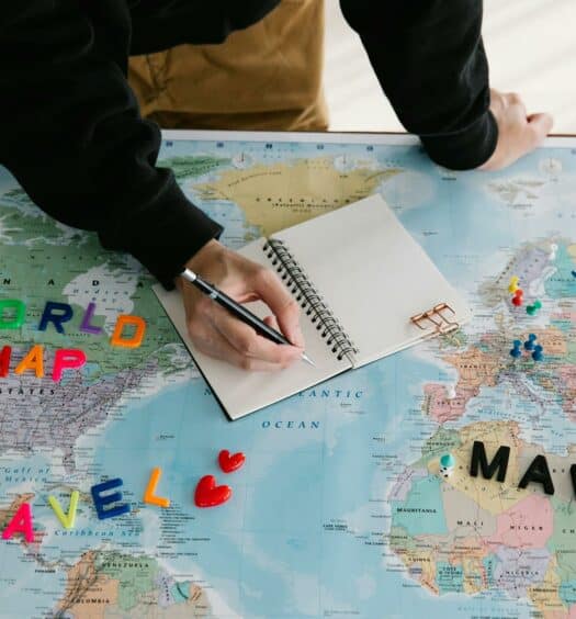 Um mapa mundi aberto com algumas peças que imitam letras formando da palavra Travel em cima dele, há também um caderno aberto e um homem escrevendo nele, para representar quanto custa um seguro viagem
