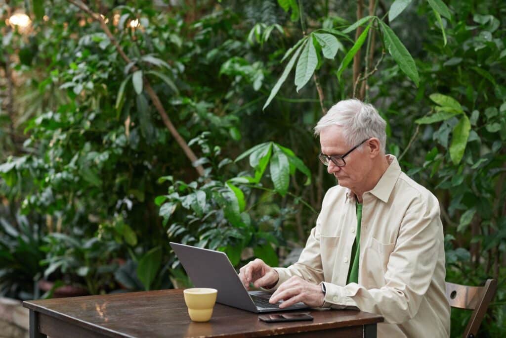 Um senhor sentado em um espaço aberto com muitas plantas ao redor, na mesa está um notebook aberto, um celular e um copo, ele está digitando no notebook, para representar seguro viagem para idosos