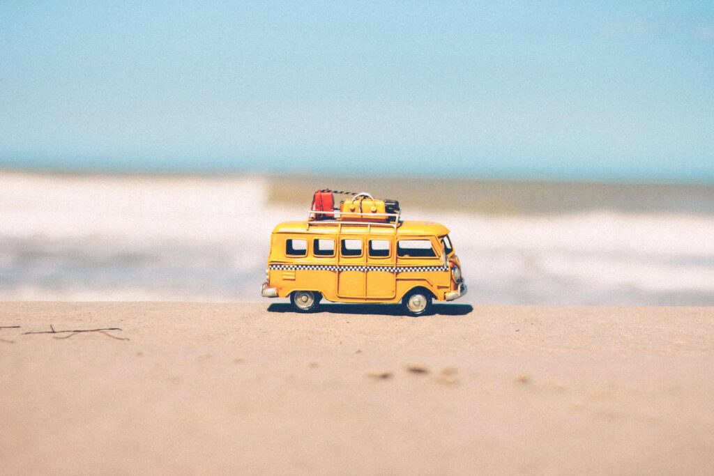 Uma kombi amarela em brinquedo com algumas malas presas no topo dela, a miniatura está na areia de uma praia com o mar ao fundo, para representar contratar seguro viagem é obrigatório