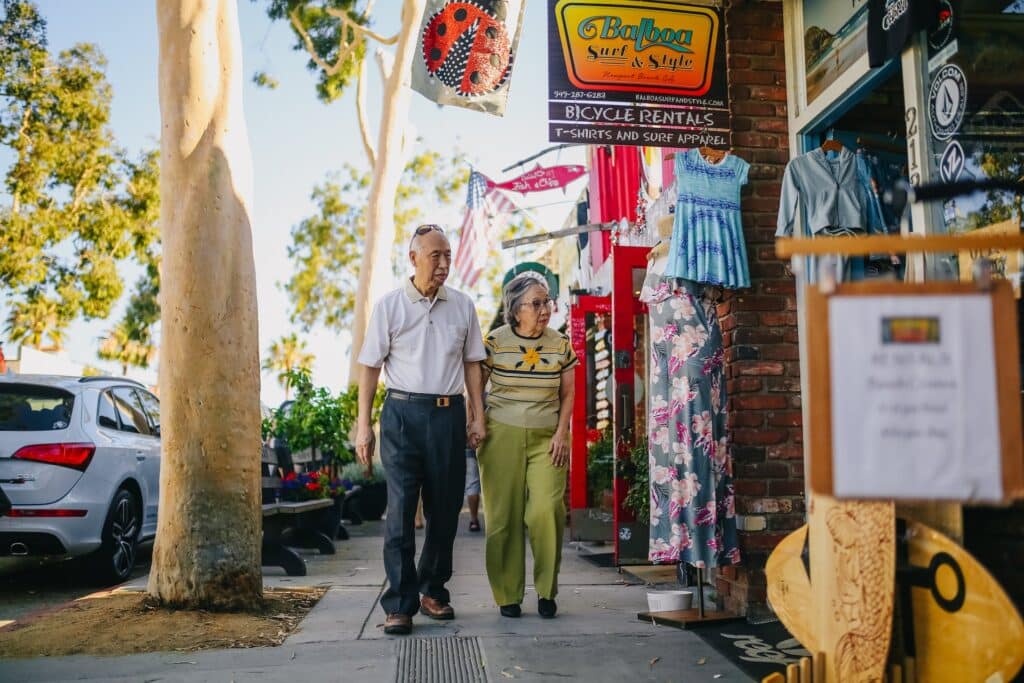 Um casal de idosos caminhando em uma rua com lojas abertas e itens pendurados na entrada do estabelecimento