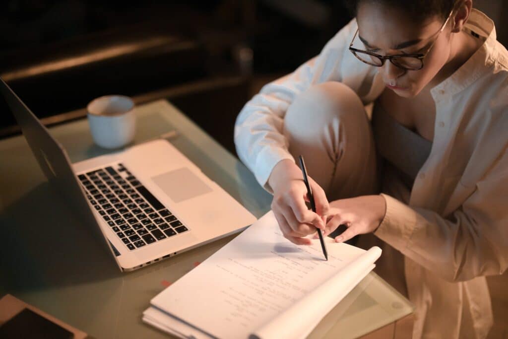 Uma mulher sentada de frente para uma mesa onde há um notebook aberto, ao lado há também um caderno onde ela está fazendo anotações usando um lápis, para representar cotação de seguro viagem