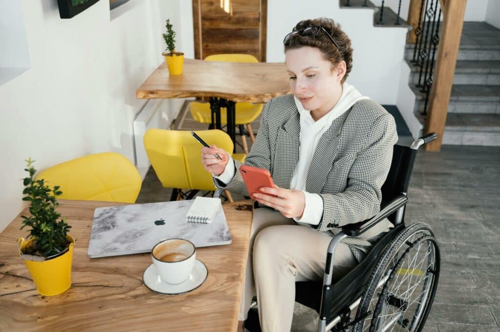 Uma mulher cadeirante sentada próxima de uma mesa de madeira, onde há um notebook, um pequeno caderno de anotações e uma xícara de café, ela está com um celular nas mãos