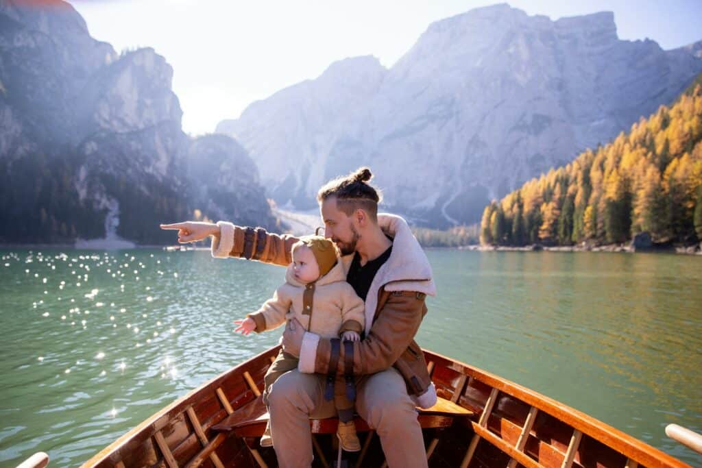 Um homem sentado em um barco em um lago límpido com muitas montanhas ao redor, em seu colo está um bebê