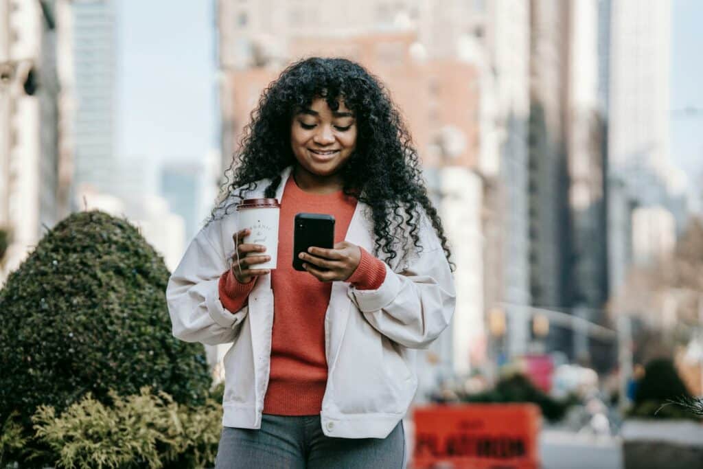 Uma mulher negra de cabelos longos e enrolados parada em uma rua movimentada, ela está segurando um celular em uma das mãos e um copo com café na outra, para representar seguro viagem Chubb