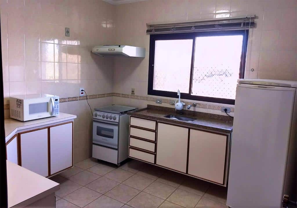 Cozinha completa em Piemonte Flat Serra Negra. Há bancadas na esquerda, e um micro-ondas está em cima de uma delas. Na direita há uma geladeira, pia e fogão.