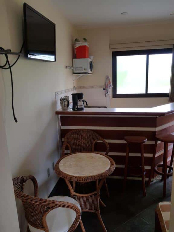 Cozinha em Piemonte Flat. No meio vemos uma mesa redonda e pequena com duas cadeiras. Atrás dela há um balcão separando a cozinha. Em cima da mesa, há uma TV fixa no alto da parede.