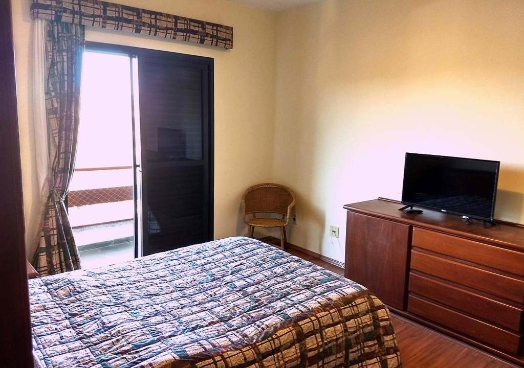 Foto do quarto em Piemonte Flat Serra Negra. A cama está no meio, vemos ela de lado. Na frente dela há uma estante de madeira com uma pequena TV em cima. Do outro lado do quarto há uma porta com varanda.
Ilustra o post sobre airbnb em Serra Negra.