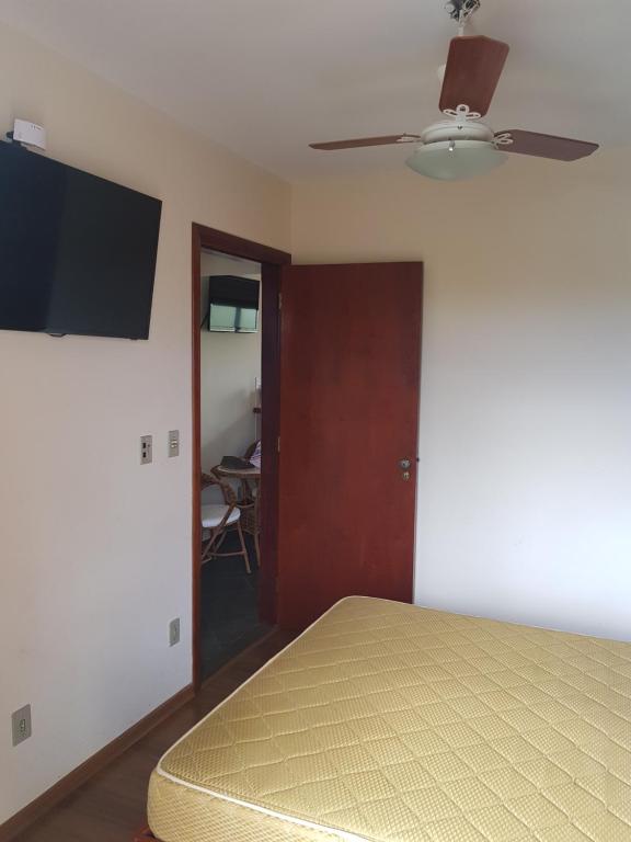Foto do quarto em Piemonte Flat. Há uma cama de casal no canto inferior direito, vemos o colchão de relance. De frente para a cama há uma TV fixa na parede. No teto há um ventilador. porta também está de frente com a cama.
Ilustra o post sobre airbnb em Serra Negra.
