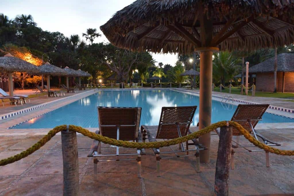 vista da piscina quadrada do Porto Preguiças Resort, em Barreirinhas, com cadeiras parecendo de palha e guarda-sol imitando coqueiros também com o topo de palha, e ao fundo, uma passagem bastante arborizada