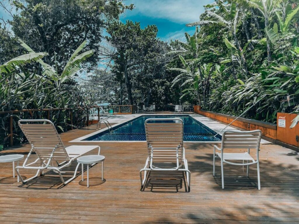 Imagem da piscina do Condomínio pé na areia – Santiago durante o dia com três cadeiras a frente e ao fundo a piscina.