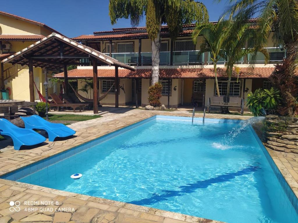 Área externa do airbnb Encantu's Flats. No lado direito da imagem está a piscina da propriedade, que é grande e retangular e no lado esquerdo estão duas espreguiçadeiras e uma parte coberta com redes de descanso. No fundo da imagem é possível ver a parte externa dos quartos.