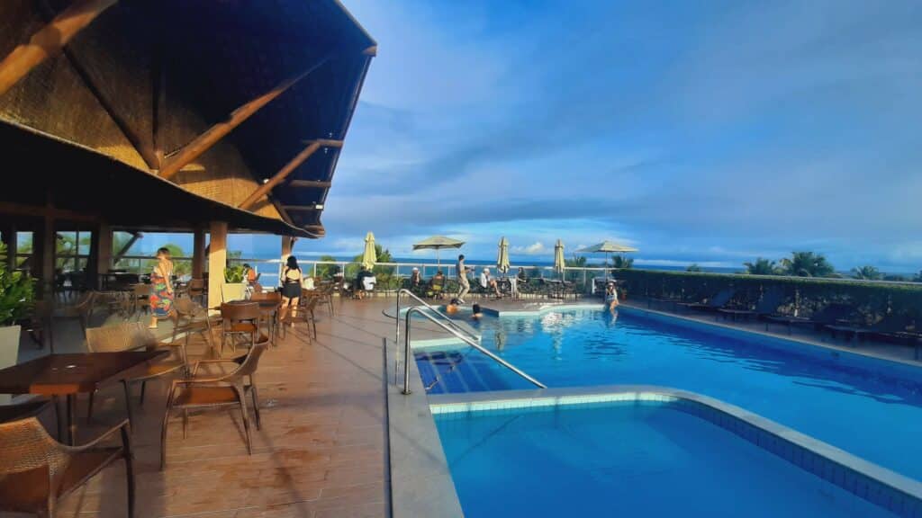 Piscina do Hotel Ponta Verde Francês, com algumas pessoas sentadas nas mesas com guarda-sóis em volta, uma menina sentada na beira da piscina e também há uma vista do mar