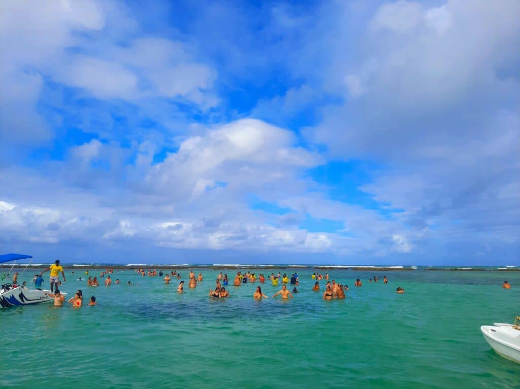 Piscina natural Ponta de Mangue, em Maragogi, com uma lancha e várias pessoas nadando em um mar verde esmeralda e cristalino, sem ondas.