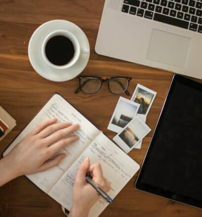 pessoa escrevendo em um planner com notebook, fotos, óculos, um cafézinho e caderno em cima de uma mesa para ilustrar o post de seguro viagem cobre cancelamento de viagem