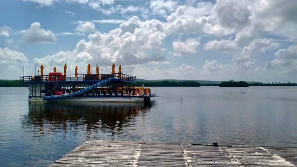 Barco grande navegando na Lagoa Mundaú, em Pontal da Barra, Maceió. O dia está ensolarado e o céu reflete nas águas do rio