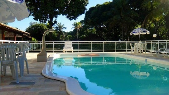 Área externa da Pousada Del Mare di Ponta Negra. Uma piscina no meio com cascata, ao redor mesas, cadeiras, guarda-sóis e árvores.