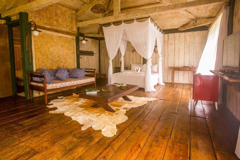 Quarto da Pousada Meu Recanto que mostra uma cama de casal encostada com a cabeceira na parede e de cada lado tem uma mesinha de madeira. Em frente a cama tem um tapete no chão com uma mesa de madeira em cima, um sofá com almofadas do lado esquerdo e do lado direito um frigobar. O chão e o teto do quarto são de madeira, imagem ilustrando post Airbnb em Ibitipoca.