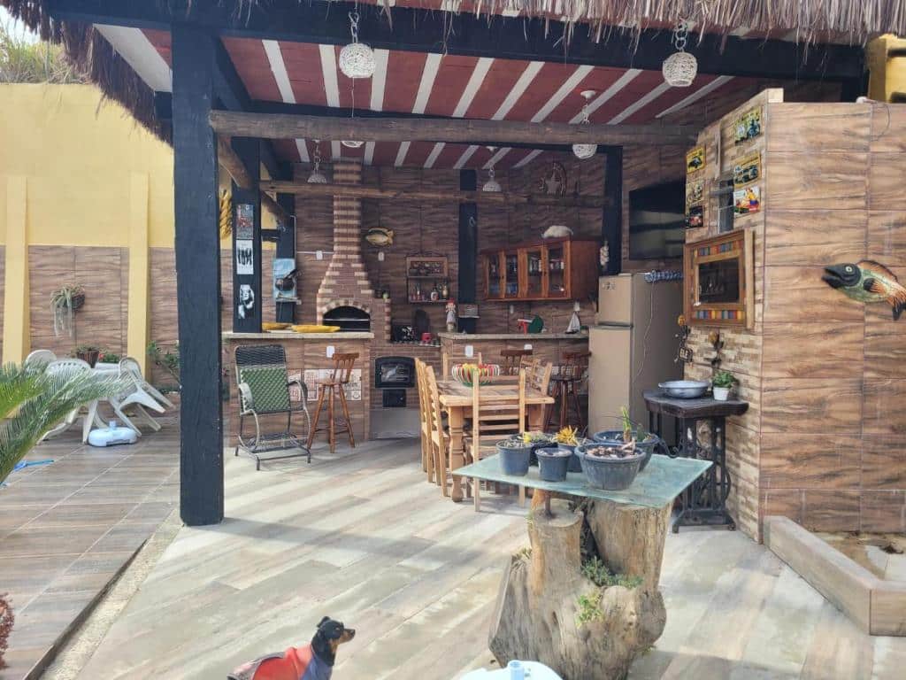 Área externa da Pousada Mirante do Pôr do Sol. No meio um tronco com vidro e vasos e um cachorrinho em baixo. No fundo uma área de lazer com churrasqueira e cozinha completa.