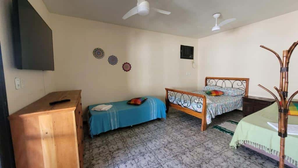 Quarto da Pousada Mirante do Pôr do Sol. Do lado direito uma cama de solteiro e uma de casal, entre elas uma cômoda, no canto direito um cabideiro. No teto dois ventiladores, no fundo, no meio uma cama de solteiro. Do lado esquerdo uma televisão e uma cômoda. Foto para ilustrar post sobre pousadas em Maricá.