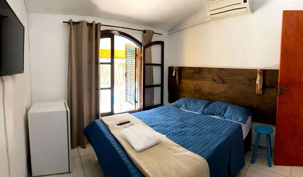 Quarto da Pousada na Praia Maricá. Uma cama de casal do lado direito com um banco de cada lado, em cima um ar-condicionado. Do lado esquerdo uma televisão e um frigobar.