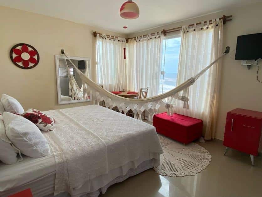 Quarto da Pousada Potigua. Uma cama de casal do lado esquerdo, de frente uma rede, uma televisão, baú, um frigobar, uma mesa com duas cadeiras e uma janela de vidro com vista para o mar. Foto para ilustrar post sobre pousadas em Maricá.