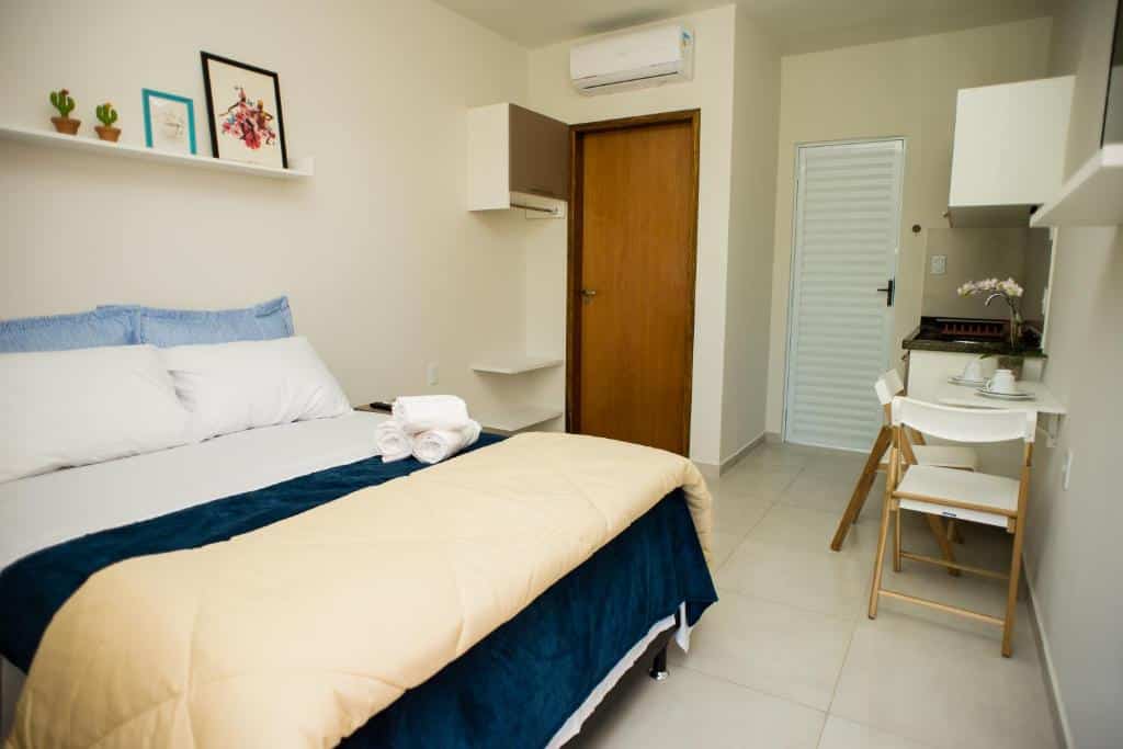 Quarto da Pousada Vila da Praia. Uma cama de casal do lado esquerdo, do lado direito uma mesa com duas cadeiras e uma mini cozinha no fundo com a porta do quarto e do lado a porta do banheiro. Foto para ilustrar post sobre pousadas em Maricá.