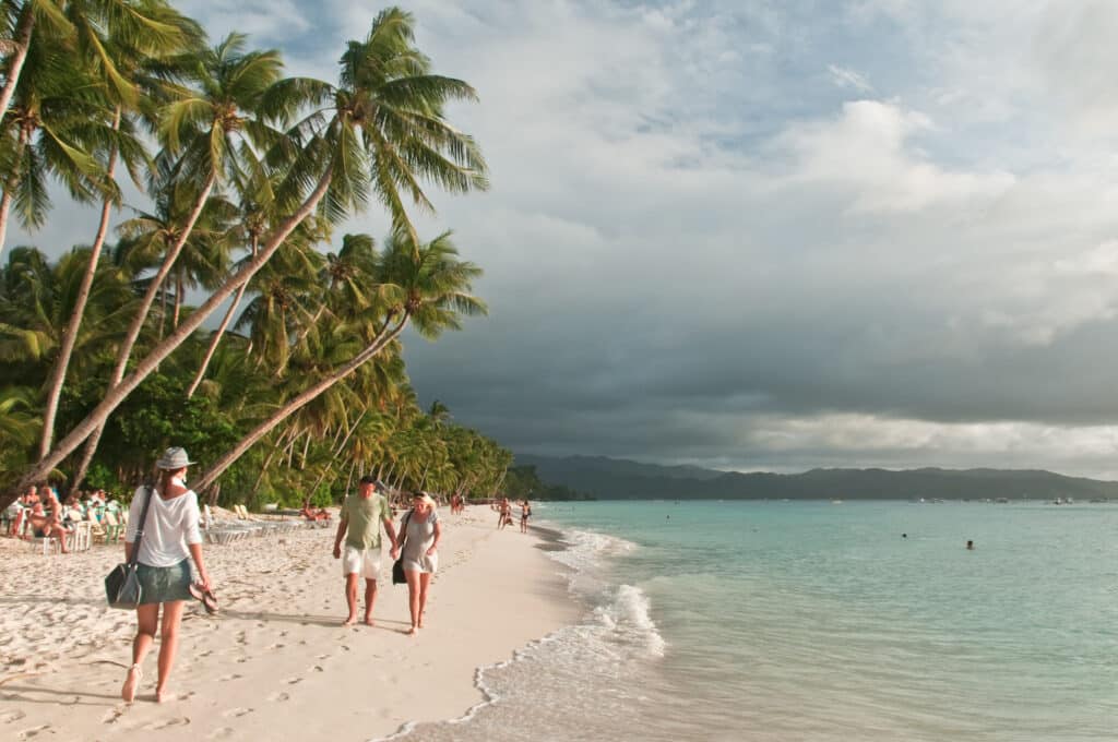 Pessoas caminhando ao longo de uma praia arenosa com palmeiras à esquerda e um oceano calmo à direita, sob um céu nublado.